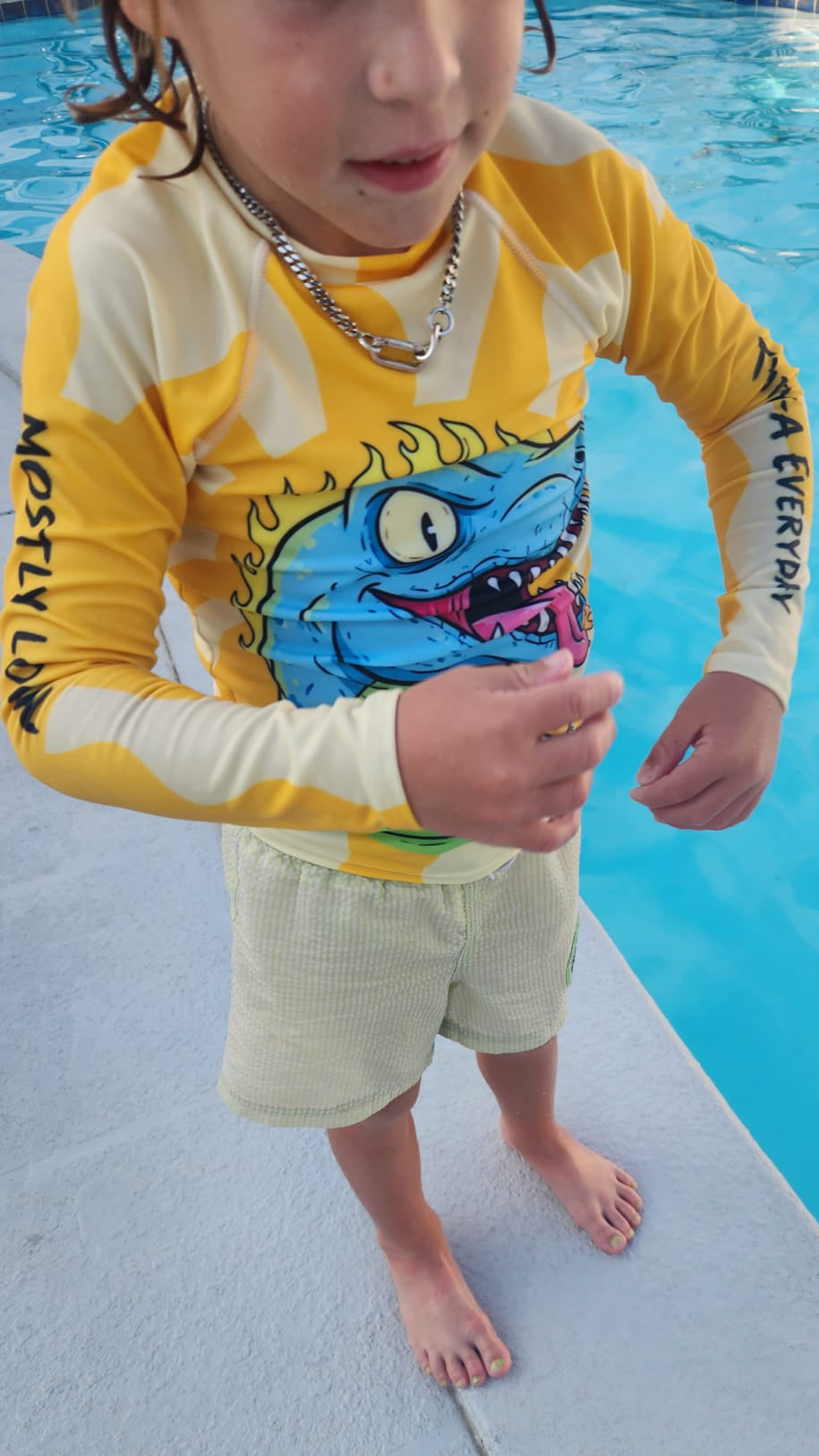 A child wearing a bright yellow and light. yellow Salt + Shine long-sleeve UPF50  swim shirt with a slobbery blue cartoon Dinosaur graphic, standing on a pool deck next to blue swimming pool water.